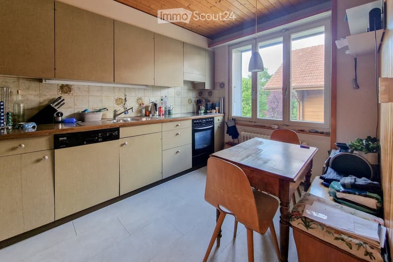 Bright kitchen with light wooden cabinets, a dining table with chairs, and various appliances including an oven and dishwasher. The space features large windows and a hanging light fixture.