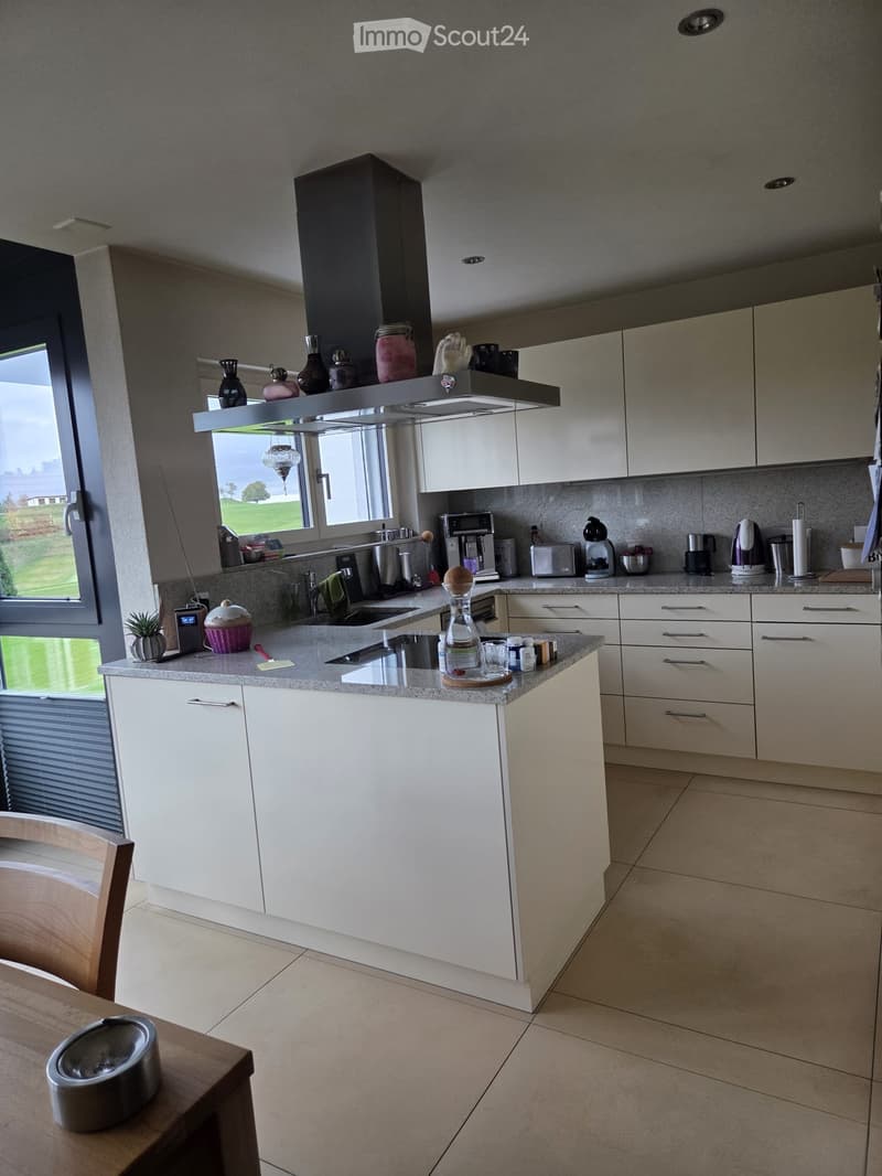 Modern kitchen with white cabinets, granite countertops, and various appliances including a coffee maker, toaster, and oven. The kitchen features a large window with a view of a green landscape.