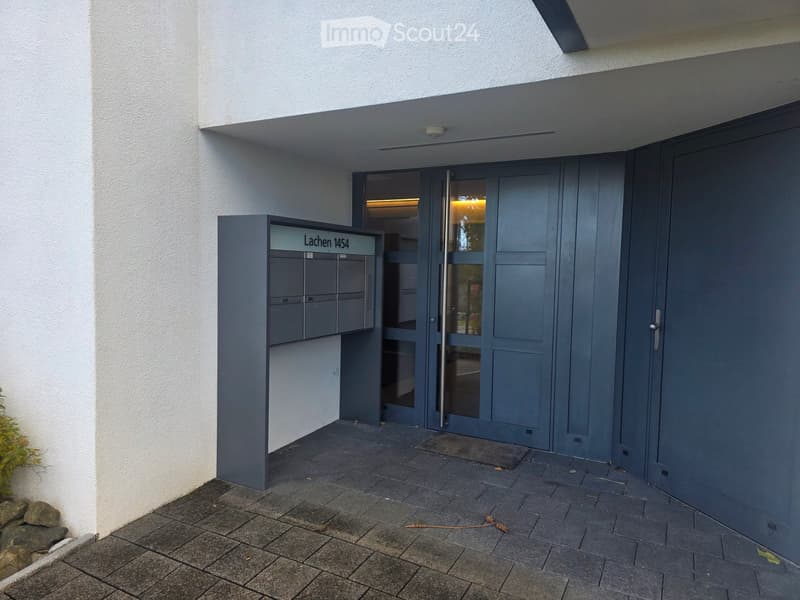 Modern building entrance with grey doors, mailboxes labeled "Lachen 1454", and a small overhang above the entrance.