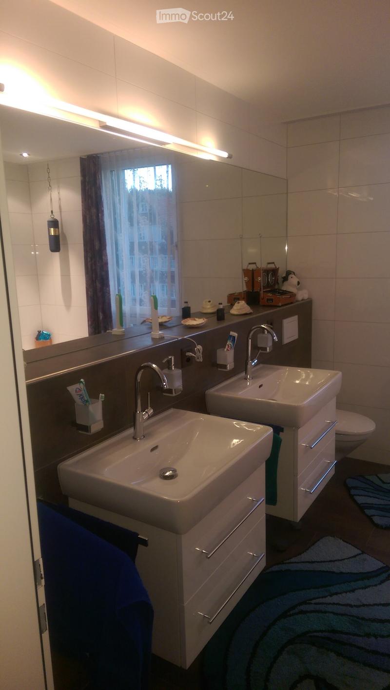 Modern bathroom with two sinks, a large mirror, cabinets, and a toilet. The space features a window with a view, a towel on the floor, and various toiletries on the counter.