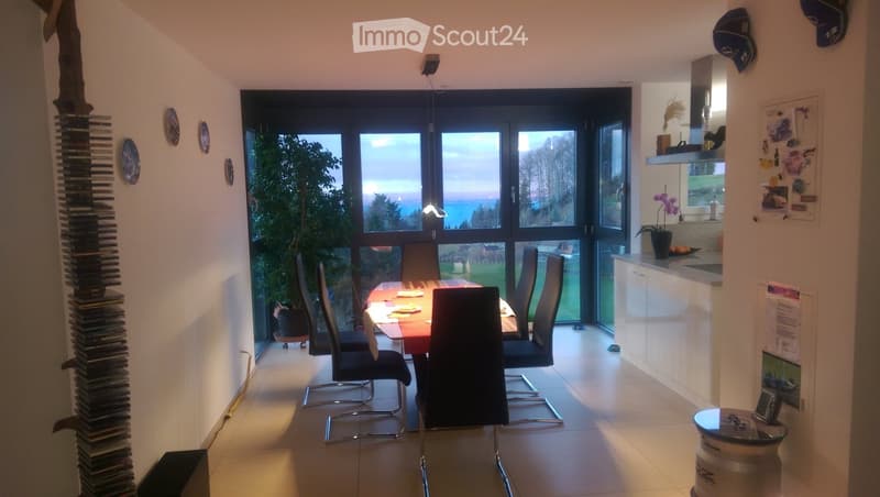 Open-plan dining and kitchen area with large windows overlooking a scenic view, featuring a dining table with chairs, a kitchen counter, and decorative elements.