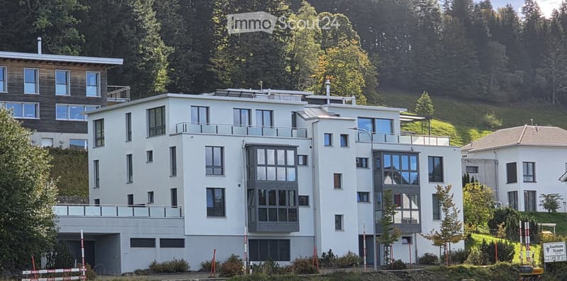 Modern multi-story residential building with white facade, large windows, and balconies, surrounded by greenery and trees.