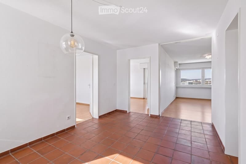 Empty hallway with white walls, tiled floor, and wooden flooring in adjacent rooms. Features a hanging glass light fixture and open doorways leading to other spaces.