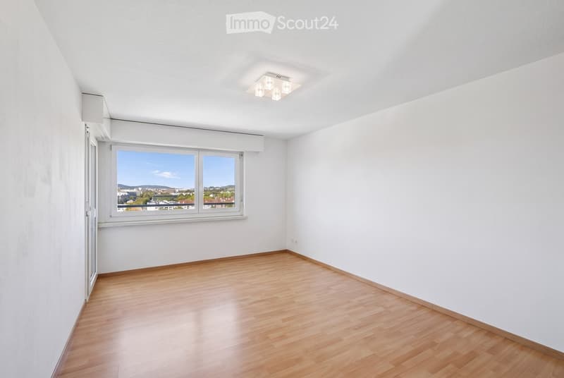 Empty room with white walls, wooden flooring, large window with a view, and a ceiling light fixture.