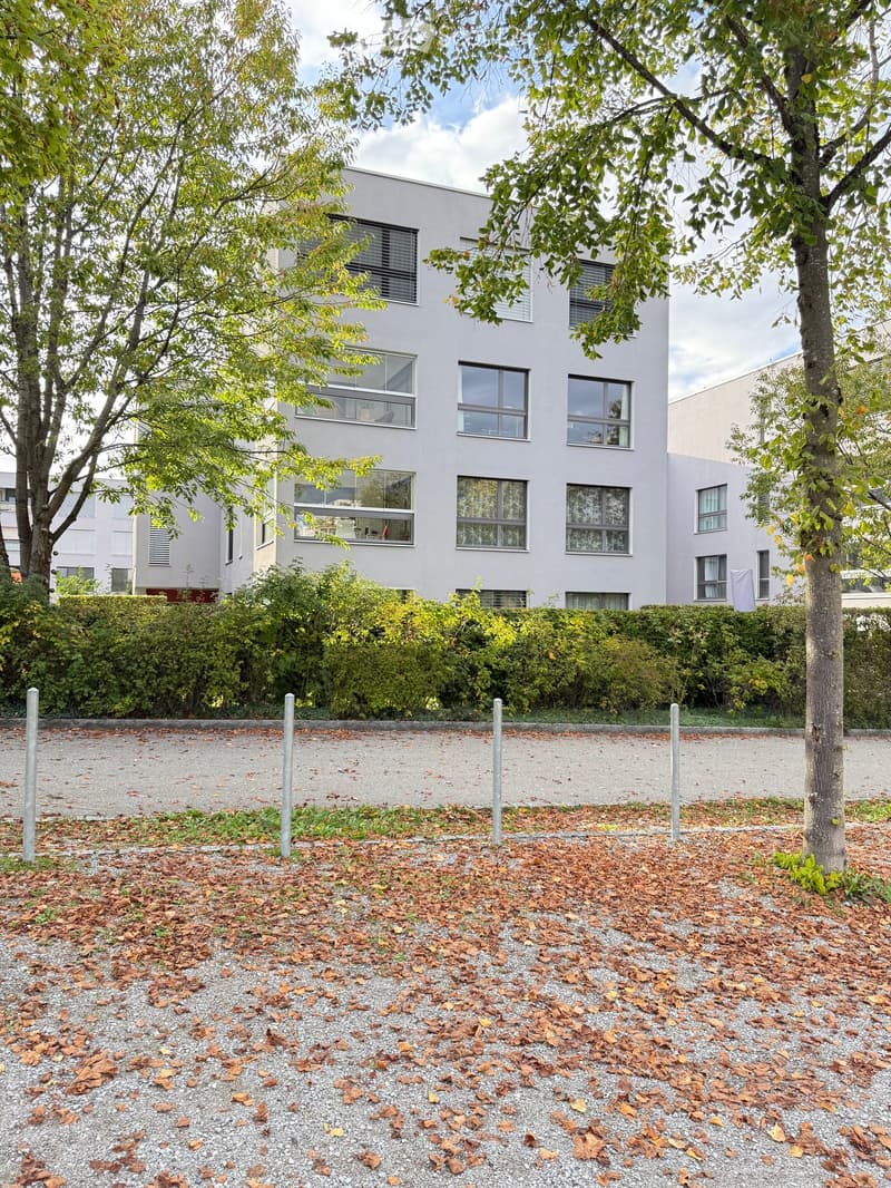 Modern two-story building with large windows and a light gray facade, surrounded by trees and shrubs.