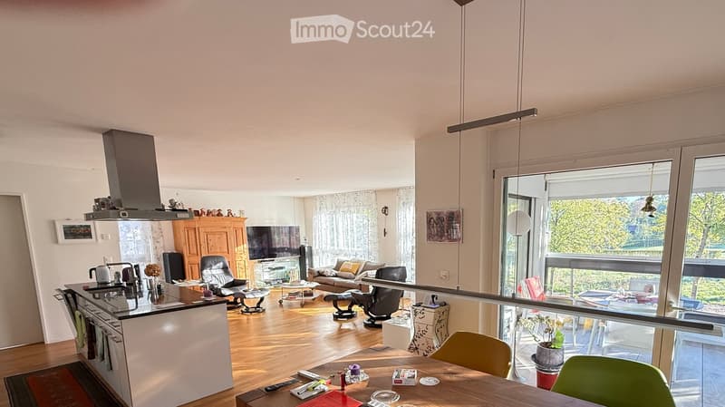 Open-plan living and dining area with modern furniture, large windows, and a view of greenery outside. Includes a sofa, coffee table, dining table with chairs, kitchen island, and various appliances.