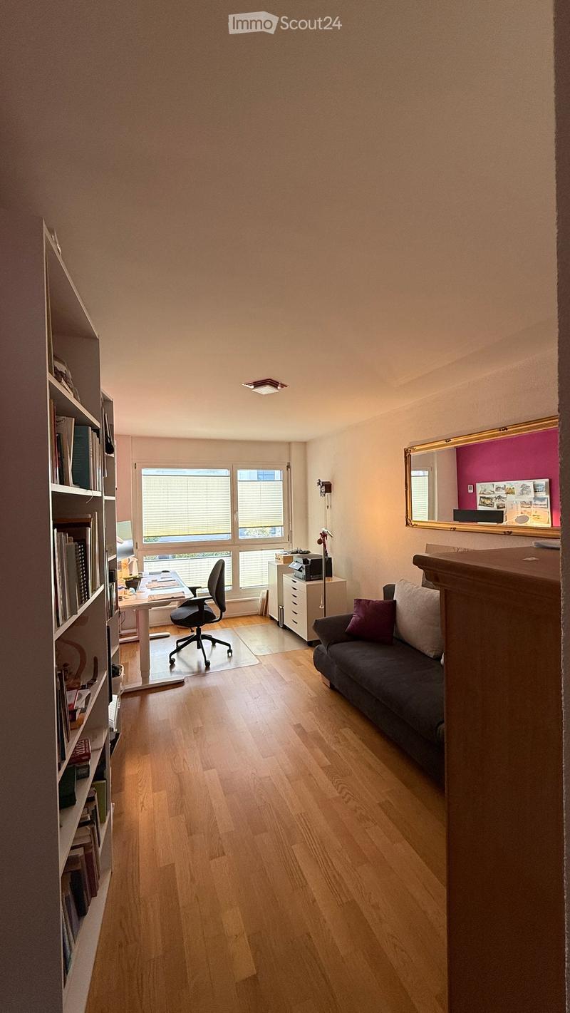 A well-lit room with wooden flooring, a sofa, a desk with a chair, a bookshelf, and a mirror on the wall.