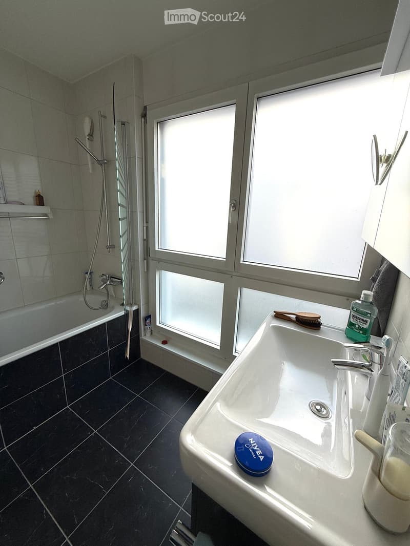 Modern bathroom with a white sink, dark tiled floor, bathtub, and shower area. The sink has a faucet, toothbrush, toothpaste, and a bottle of mouthwash. A blue Nivea container is placed on the sink.