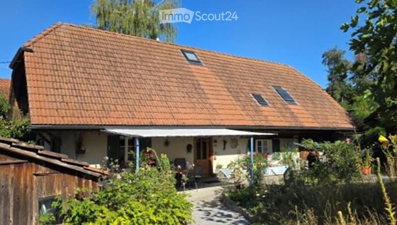 A two-story house with a red-tiled roof, white walls, and a covered patio area. The house is surrounded by a well-maintained garden with various plants and trees.