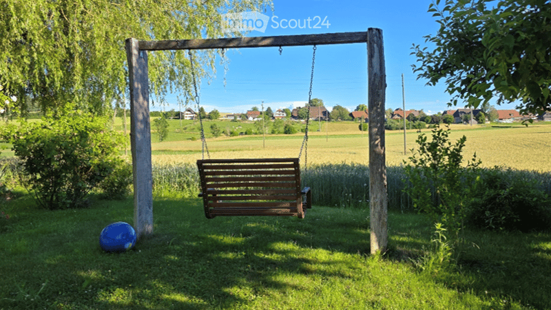 A peaceful garden area with a wooden swing, a blue ball on the grass, and a scenic view of a rural landscape with fields and houses in the distance.