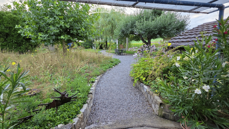 A well-maintained garden with a gravel pathway, surrounded by lush greenery, flowers, and trees. There is a small wooden structure and a bench visible in the background.