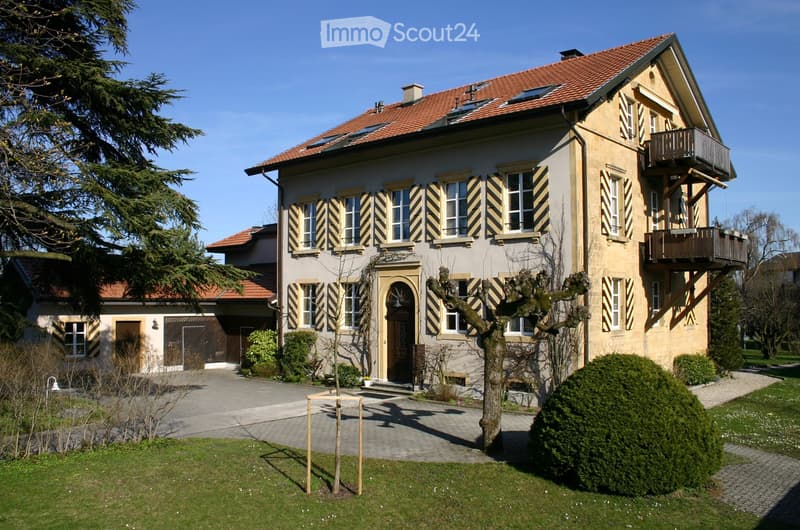 Two-story house with red-tiled roof, beige facade, multiple windows with shutters, and a balcony on the upper floor. The house is surrounded by a well-maintained garden with trees and shrubs.