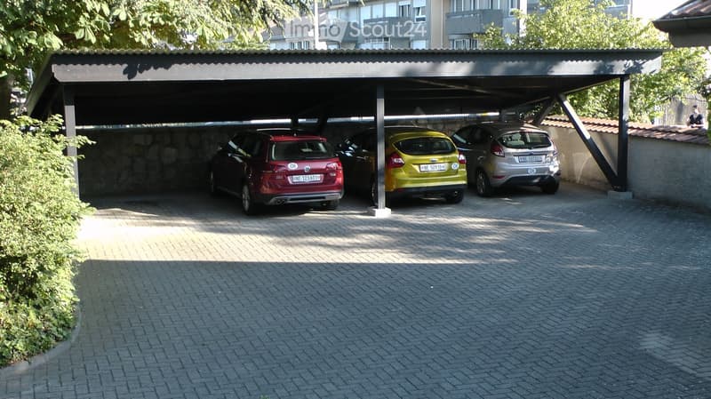 Carport with three parked cars under a covered structure, surrounded by greenery and a paved area.