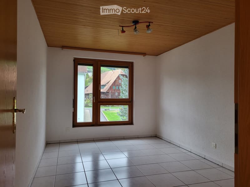 Empty room with white tiled floor, light-colored walls, and a large window with wooden frame offering a view of the outside. The ceiling is wooden with built-in lighting fixtures.
