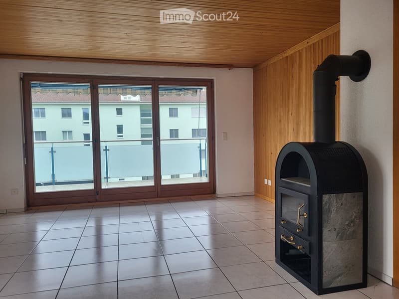 Modern room with wooden ceiling, tiled floor, large window with balcony view, and a black wood-burning stove.