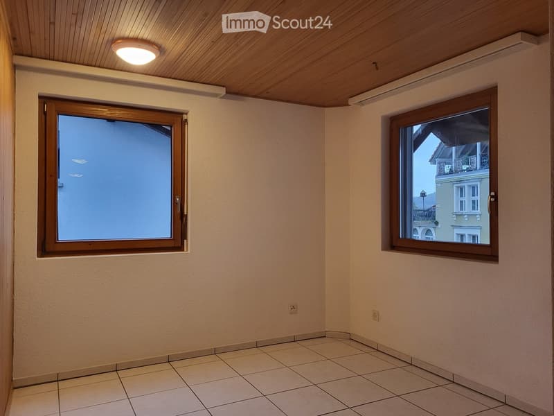 Empty room with tiled floor, light-colored walls, and two windows with wooden frames. The room has a recessed ceiling light and visible electrical outlets.