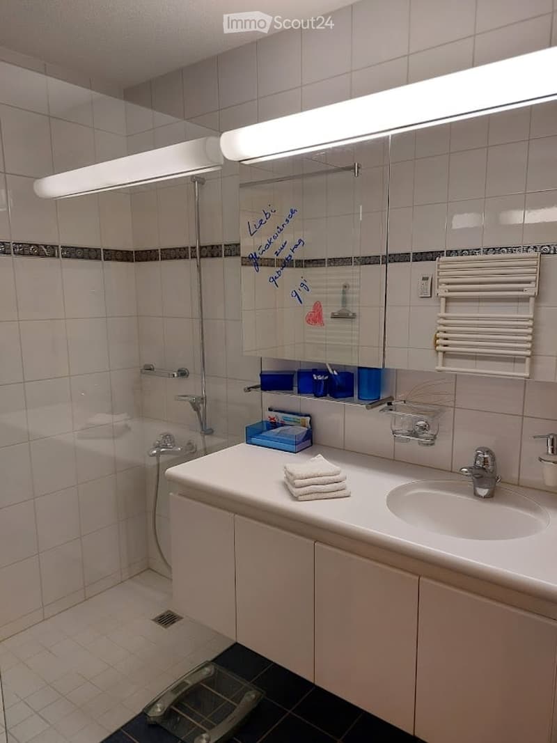 Modern bathroom with white tiles, a large mirror, a sink with a faucet, a shower area, a bathtub, and a radiator. There are towels and cups on the sink counter.