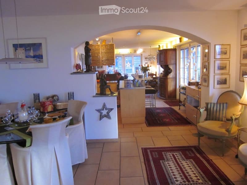 A well-lit living and dining area with a dining table, chairs, a kitchen area with a counter, stools, and a lamp. The space features a mix of modern and traditional decor, including framed pictures and a patterned rug.