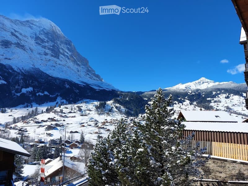 Snow-covered village with traditional wooden houses, surrounded by snow-capped mountains under a clear blue sky.