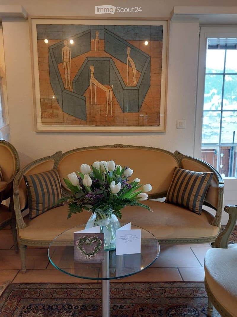 Elegant living room with a cream-colored sofa, glass coffee table, decorative flower arrangement, and artwork on the wall. The room features large windows with a scenic view.