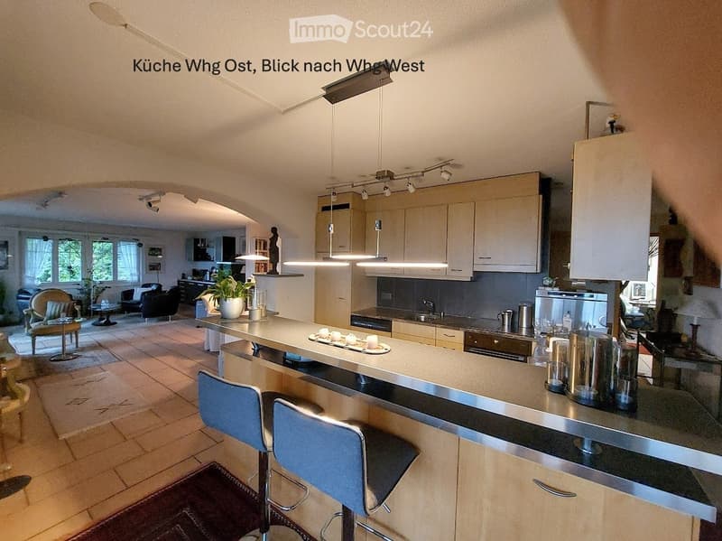 Modern kitchen with light-colored cabinets, a central island with bar stools, and a living area visible in the background with seating and a coffee table.