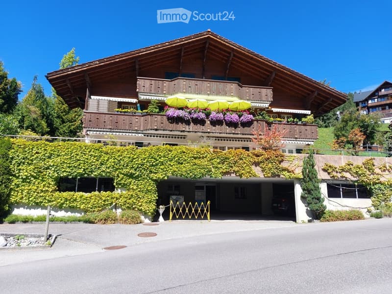 A large, two-story wooden house with a steeply pitched roof, balconies adorned with colorful flower boxes, and a covered parking area partially covered by climbing plants.