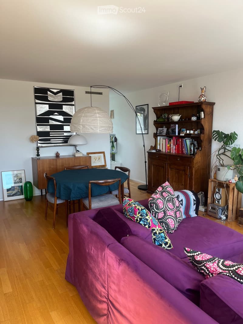 A modern living room with a purple sofa, colorful patterned pillows, a wooden dining table with chairs, a bookshelf, and decorative items.