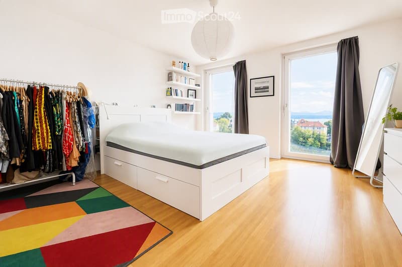 Modern bedroom with a white platform bed, colorful geometric rug, large window with view, wardrobe, shelves, and a hanging lamp.