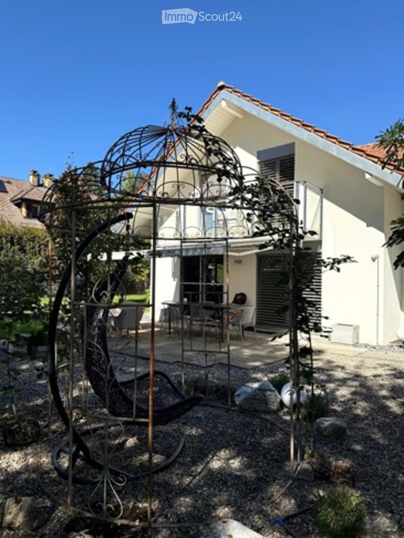 A modern house with a balcony and a decorative metal gazebo in the garden area. The garden features gravel ground, a few potted plants, and a comfortable outdoor seating area.
