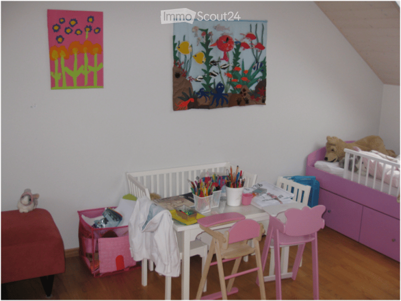 A cozy children's room with a crib, a small desk with chairs, a toy box, and a bed with stuffed animals. The room features colorful artwork on the walls and a wooden floor.