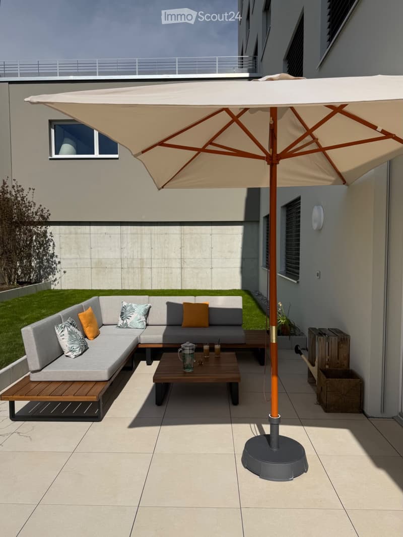 Modern outdoor patio with a grey sectional sofa, wooden coffee table, and large umbrella providing shade.