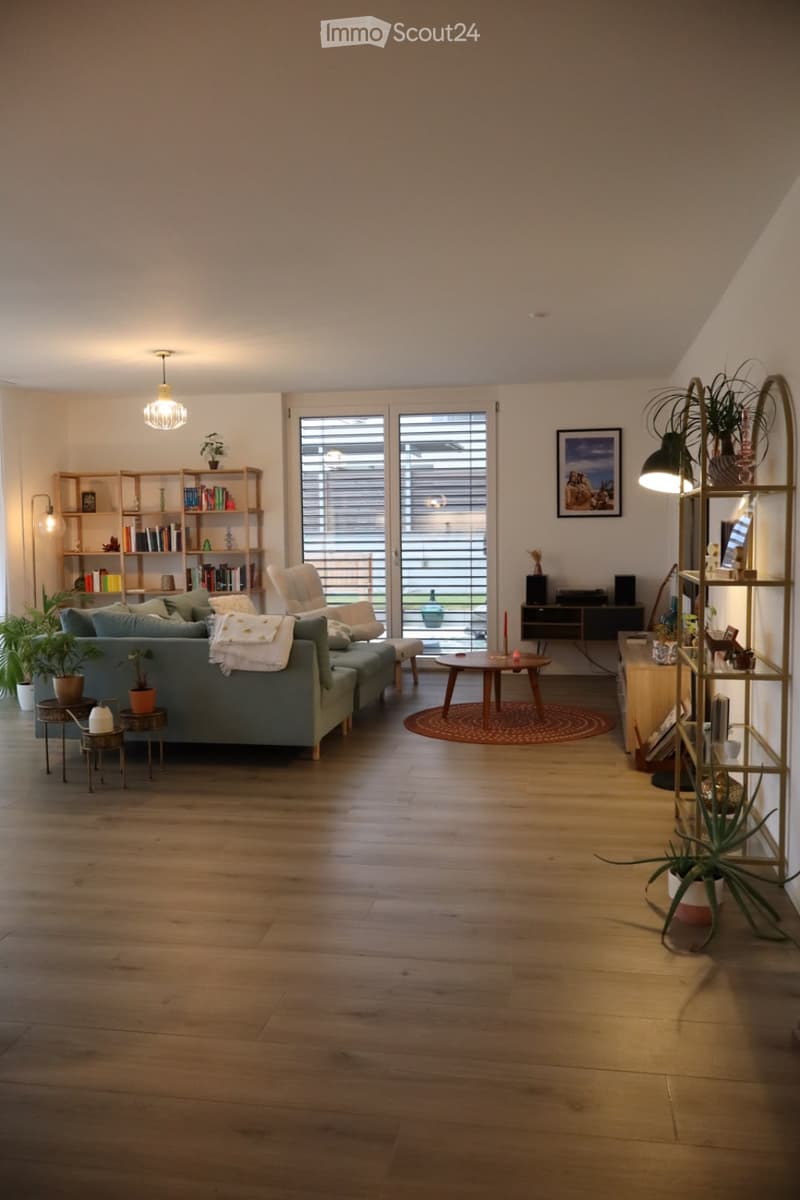 Modern living room with light wooden flooring, large windows, a grey sofa, coffee table, bookshelf, and decorative plants.