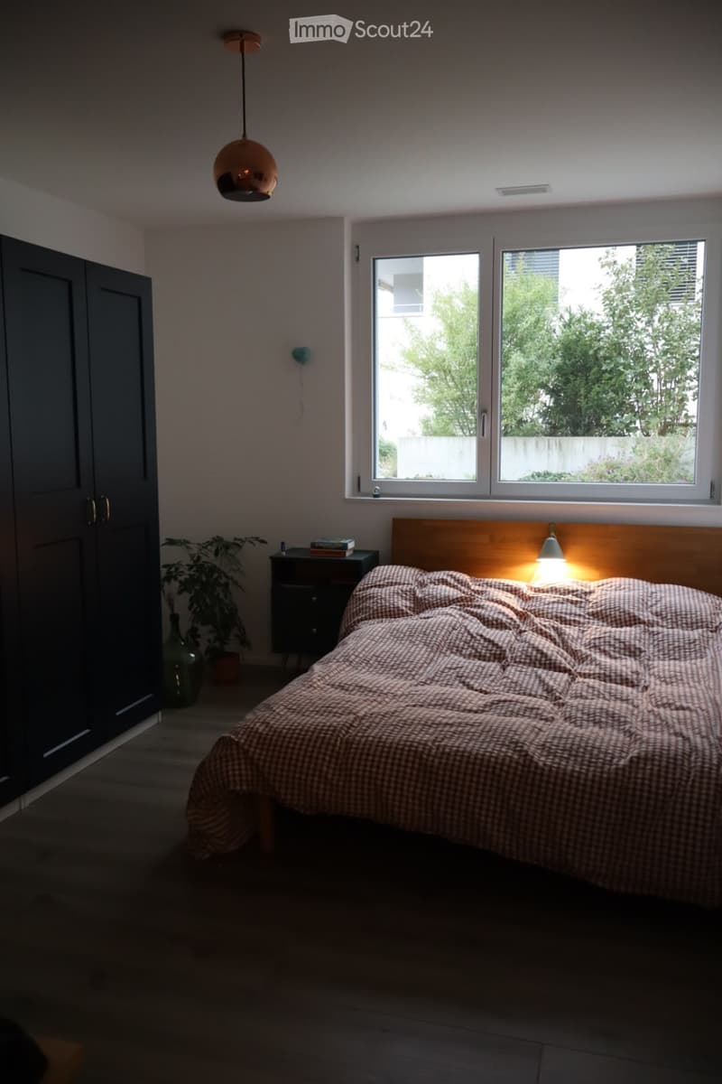 Modern bedroom with a large bed, nightstand, lamp, plant, and wardrobe. Large windows provide natural light.