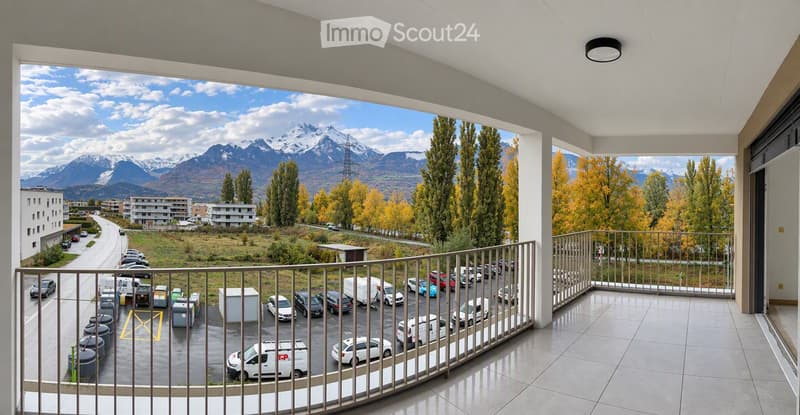 Balcony with a view of a parking lot, road, and distant mountains. The balcony has a metal railing and tiled flooring.