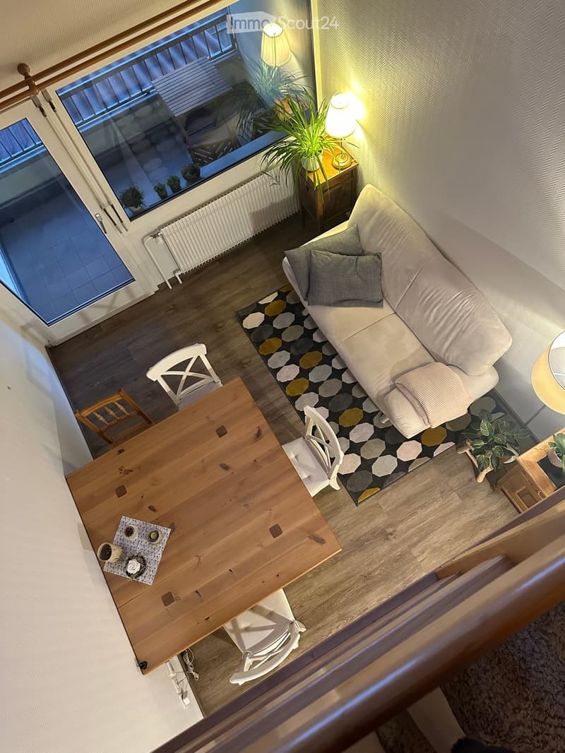 A modern living room with a white sofa, wooden dining table, white chairs, a patterned rug, and a side table with a lamp and plant. The room features large windows with a view of another room.