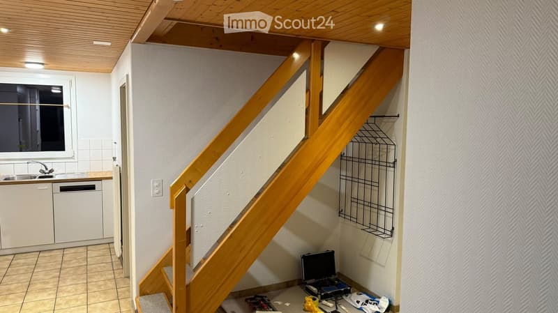A narrow hallway with a wooden staircase leading upwards, white walls, and a small kitchen area visible to the left with a sink, dishwasher, and cabinets.
