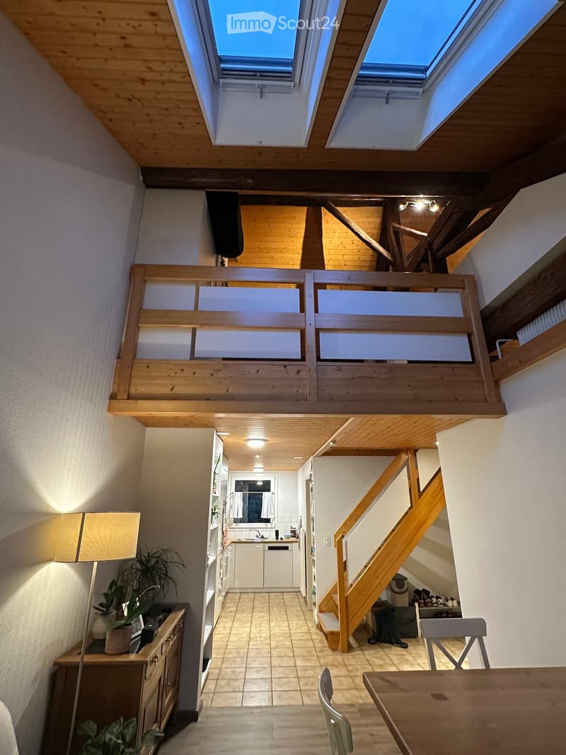 The image shows a modern, well-lit interior space with wooden beams and skylights. The main area includes a dining table with chairs, a lamp, and a cabinet with a potted plant. A staircase leads to an upper level, and a kitchen area is visible with white