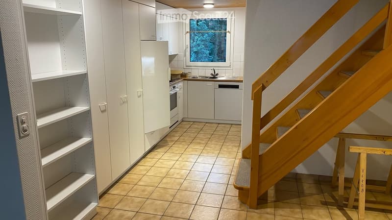 Empty kitchen with white cabinets, tiled floor, and a window overlooking a forested area. A wooden staircase leads upwards.