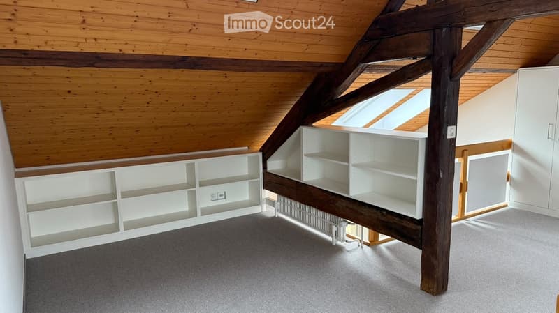 An attic room with wooden beams, built-in white shelving units, and skylight windows allowing natural light to enter.