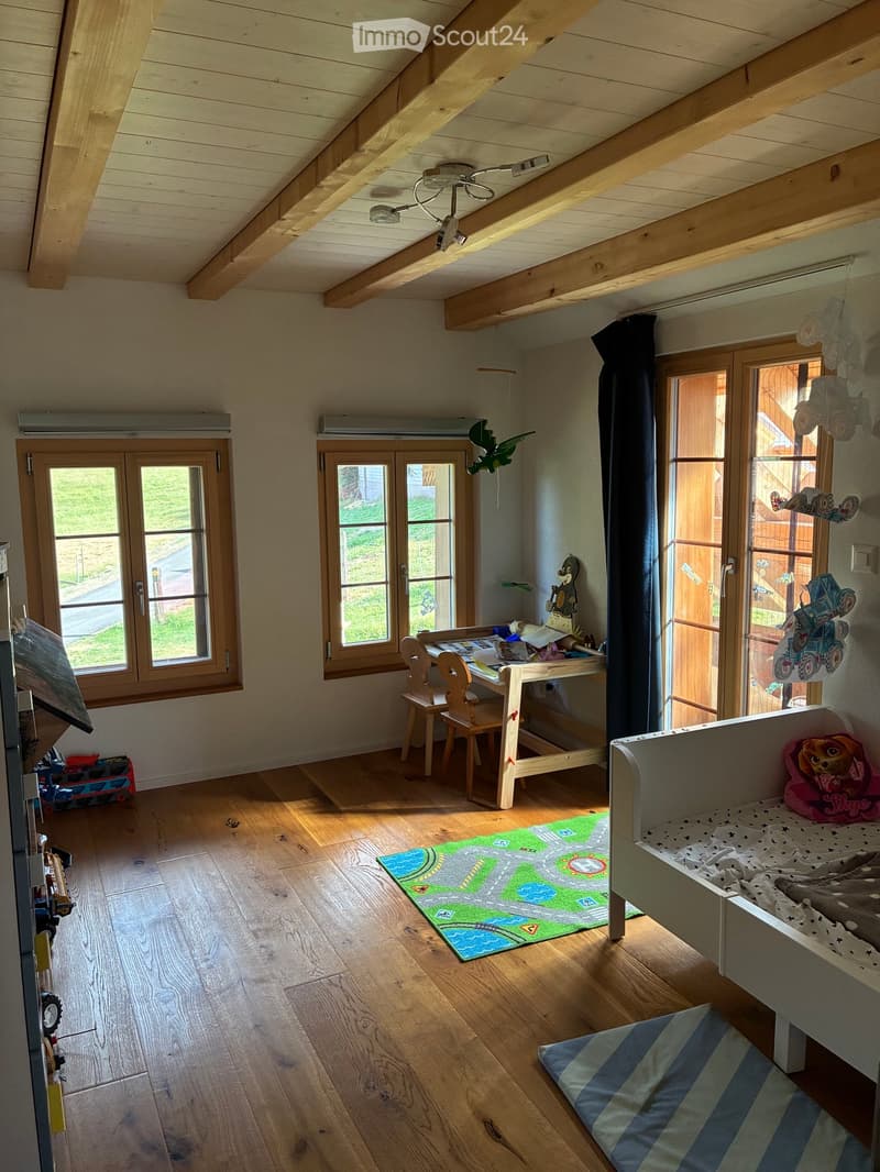Brightly lit children's room with wooden floors, large windows, a desk with chairs, a toy bed, and various toys including a rug, pillows, and blankets.