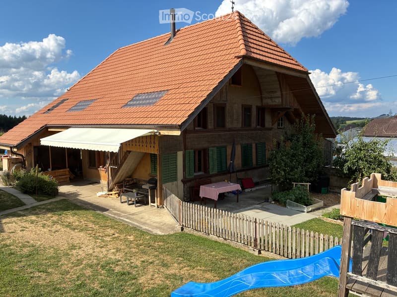 Two-story house with red-tiled roof, green shutters, covered patio with outdoor furniture, and a small garden with a slide.