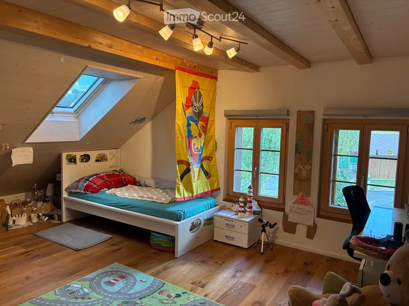 A cozy attic bedroom with wooden beams, skylight, and large windows. Features a bed with colorful bedding, a nightstand with a lamp, a small table with a telescope, and a colorful rug with a race track design.