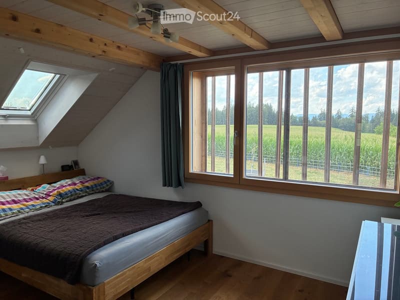 Cozy attic bedroom with wooden beams, large windows overlooking a field, a bed with a colorful quilt, and a small table lamp.