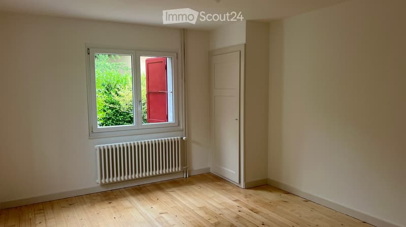 Empty room with wooden flooring, white walls, a radiator, and a window showing greenery outside.