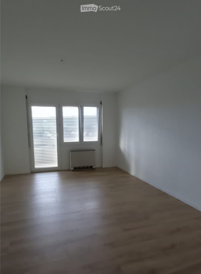 Empty room with wooden flooring, white walls, and a radiator under a window with blinds.