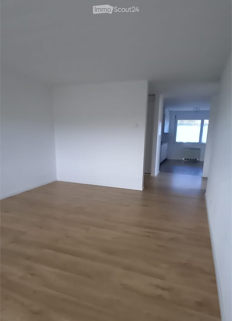 Empty hallway with wooden flooring and white walls, leading to another room with a window and radiator.