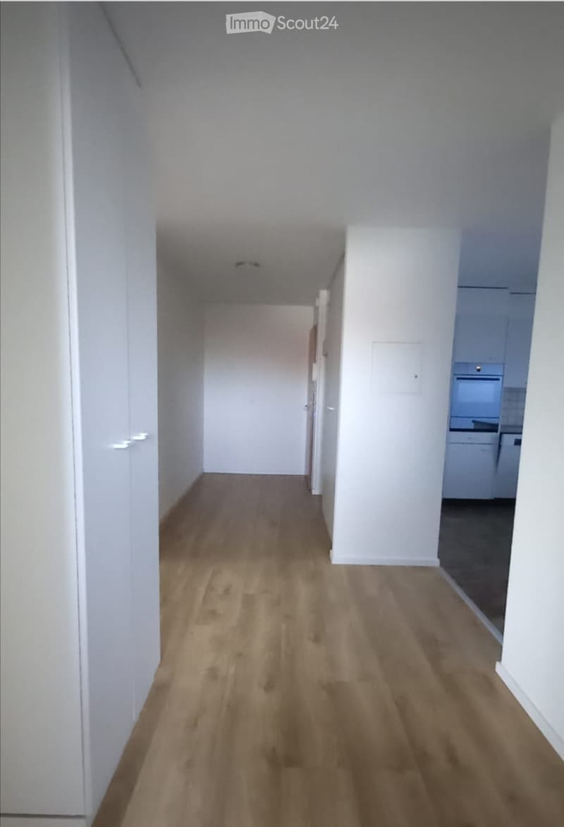 Empty hallway with light wooden flooring and white walls, leading to a partially visible kitchen area.