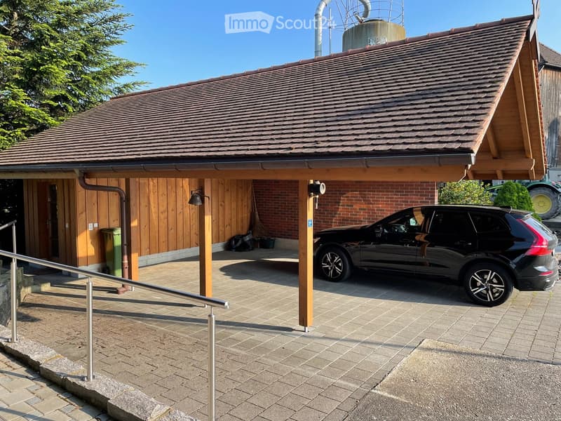 Carport with a black SUV parked underneath, wooden walls, tiled roof, and a paved entrance area with a handrail.