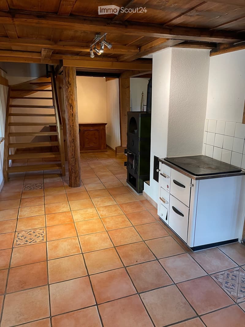 The room features a wooden staircase leading upwards, a central wooden pillar, a wooden cabinet, a wooden stove, and a kitchen area with white cabinets and an induction cooktop. The floor is tiled with terracotta tiles.