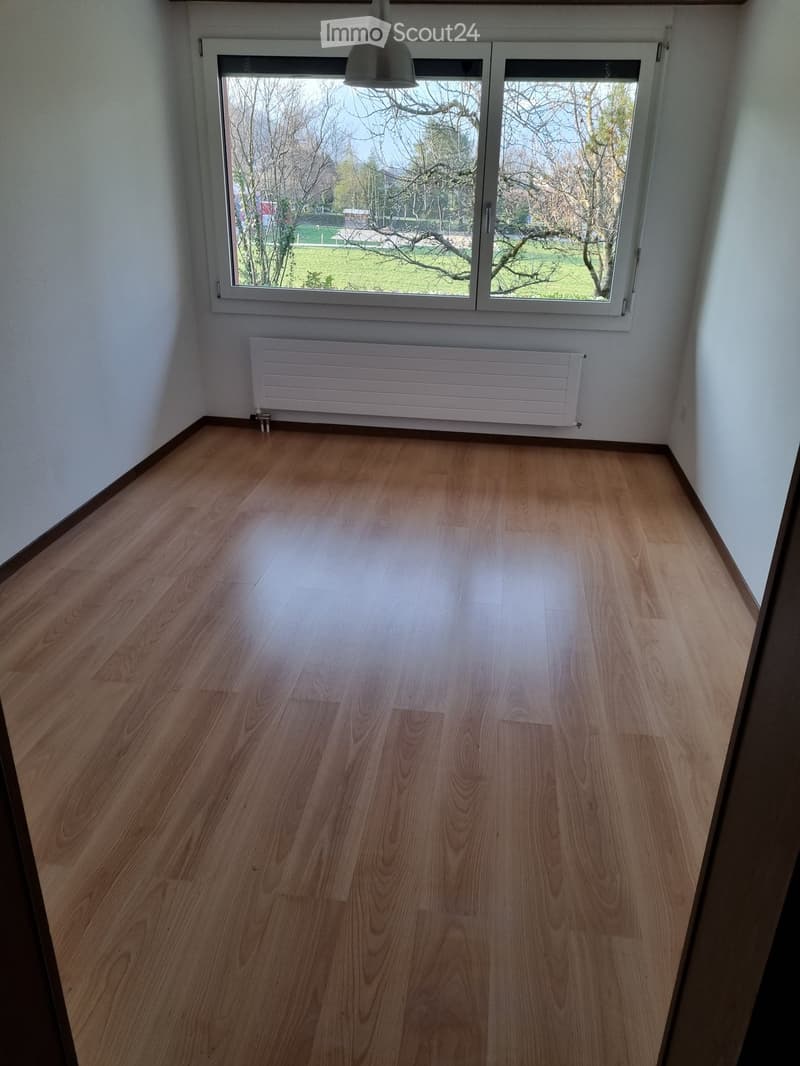 Empty room with light wooden flooring, white walls, a radiator, and a large window offering a view of a green outdoor area.